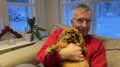 En man med en hund i famnen sittter i en soffa i ett julpyntat vardagsrum med snöiga fönster bakom sig. 