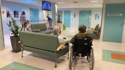 Personer i väntrummet vid en hälsocentral, en person i rullstol längst fram. 