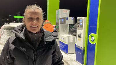 En kvinna med svart jacka står vid en tankstation med en snötäckt bil i bakgrunden. 