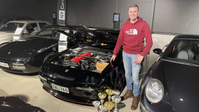En man står bredvid en svart bil som har motorhuven uppe. På golvet intill bilen ligger också en mängd pokaler.