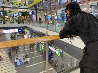 En man med långt, svart hår i småflätor och svart mössa lutar sig mot ett räcke i köpcentermiljö.