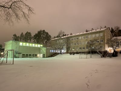 En 1950-talsskola i Karis, grå, rappad, skymning. Lite snö på marken. Belysning i en del av byggnaden. 