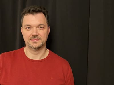 En medelålders man iklädd röd tröja tittar in i kameran - svart draperi i bakgrunden.