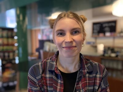 Personporträtt. Ung kvinna i rutig skjorta står i cafémiljö, ler och ser in i kameran.
