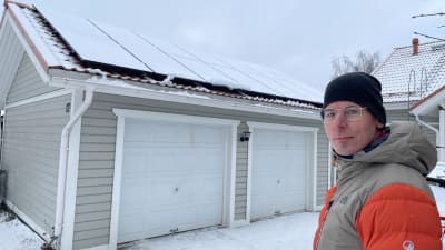 En man med glasögon och mössa står vid ett snötäckt garage med solpaneler. 