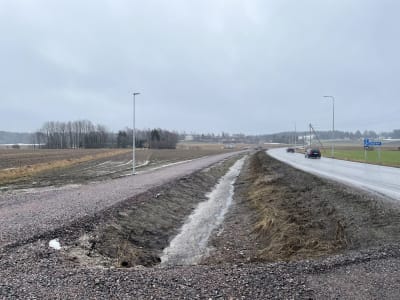 En asfalterad väg till höger där det kör några bilar. Till vänster en cykelväg som håller på att byggas, den är ännu oasfalterad.