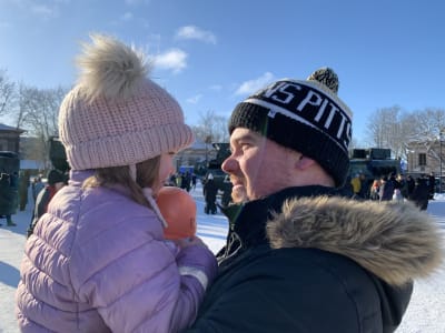 Profilerna av en man och hans dotter, flickan upplyft i mannens famn, båda vinterklädda med mössor på huvudet.