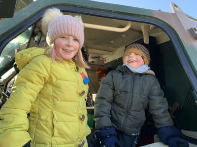 Två barn, en flicka och en pojke, i vinterkläder har klättrat upp i förarhytten på ett trupptransportfordon och ler stort.