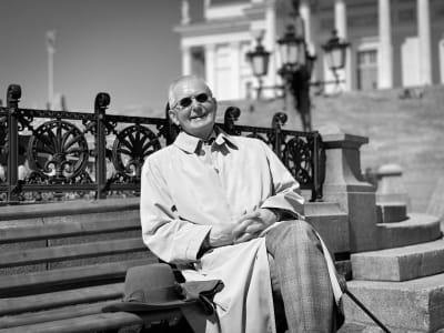Svartvit bild på Matti Klinge som sitter på en bänk på torget framför domkyrkan i Helsingfors, leende och klädd i solglasögon. 