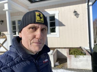 En äldre man med mössa på  huvudet står framför ett hus, tittar rakt in i kameran. Solsken.