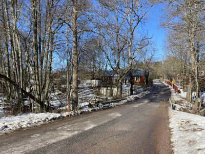 En liten byväg, asfalterad, snö på vägrenarna. Röda hus och mitt på bilden en bro över en bäck som knappt syns. Högra bara träd. Längst borta syns en plankorsning.