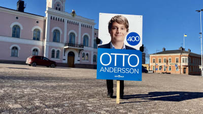 En person som inte syns håller upp en skylt med en valaffisch på Otto Andersson. Personen med skylten står på Lovisa torg och det rosa stadshuset syns i bakgrunden.