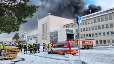 Brandmän och brandbilar intill branddrabbad byggnad på industriområde. I bakgrunden syns stort mörkt rökmoln.