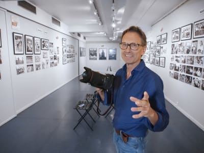 En man håller en systemkamera i handen, i bakgrunden väggar täckta med fotografier
