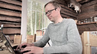 En man med stickad tröja och glasögon sitter vid ett bord med en bärbar dator