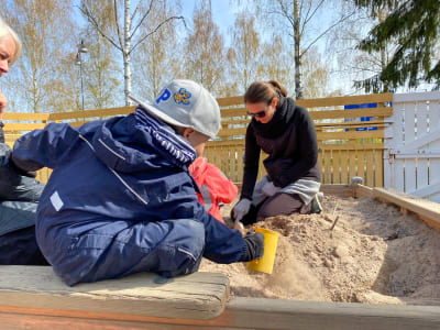 En kvinna gräver med liten spade i sandlåda. Framför henne till vänster syns en ung pojke bakifrån, han gräver också i lådan.