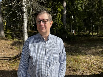 En man i rutig skjorta och glasögon står framför ett skogsparti. 
