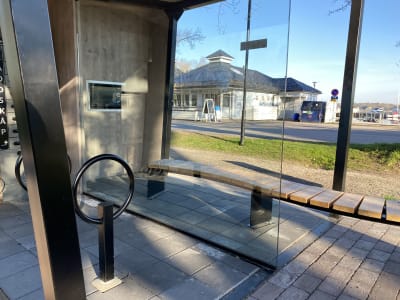 En bänk på stenplattor vid en vägg i trä. På väggen en elektronisk skärm. Ett ställ för elcyklar. I bakgrunden en glasskiosk och restautang och båtar.