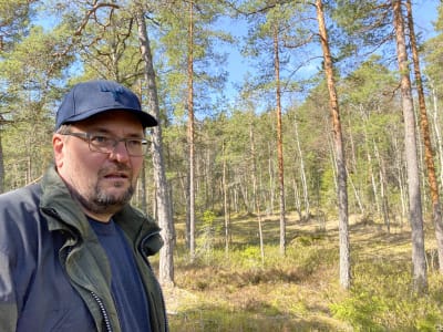 Johnny Sved, ansvarig för skogsbruksutbildningen vid YH Novia i Raseborg. En medelålders man i skärmmössa med glasögon och  lite skägg. I bakgrunden skymtar skog och blå himmel. Solsken.