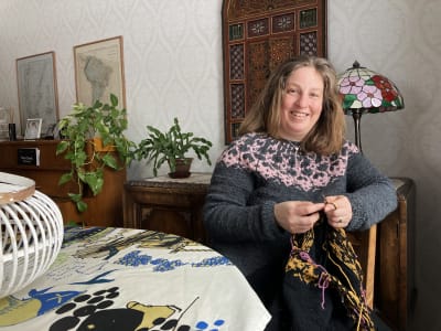Maria Ehrnström-Fuentes sitter hemma i sitt vardagsrum och visar upp sin Strömsötröja och den nya tröjan hon stickar på.