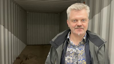En man med grågrön jacka och blommig skjorta står i öppningen av en stor kontainer. 