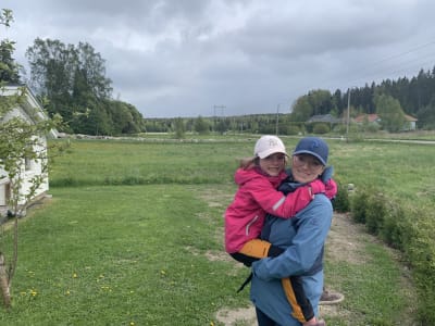 Personporträtt på en kvinna med ett barn i famnen. I bakgrunden grönskande ängar och en grusväg.