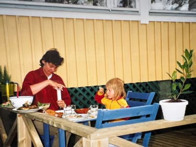 En kvinna och en liten flicka sitter vid ett matbord utomhus i solen intill en sommarstuga.