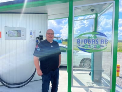 En leende man står framför en tankstation för biogas. Bakom honom står en vit personbil.