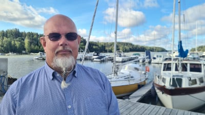 En man med vitt skägg står på en brygga. I bakgrunden segelbåtar. 
