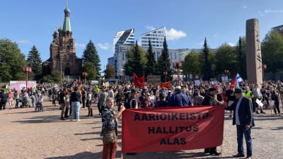Ihmisiä on kokoontunut mielenosoitukseen aukiolle. Etualalla on punainen lippu, jossa lukee äärioikeistohallitus alas.