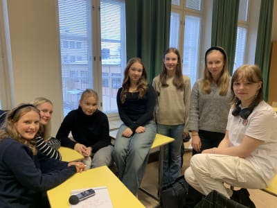 En grupp på sju skolelever, samtliga flickor i tonåren, sitter tillsammans i klassrum och tittar in i kameran.