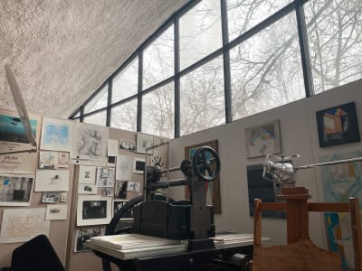 Ett arbetshörn som innehåller en gammaldags grafikpress, på väggarna i bakgrunden finns upphängd konst och grafik samt stora fönster.