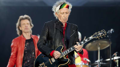 På bilden syns rockbandet Rolling Stones uppträda under en konsert i Berlin år 2018. I förgrunden syns gitarristen Keith Richards iklädd svarta kläder och en färggrann bandana.