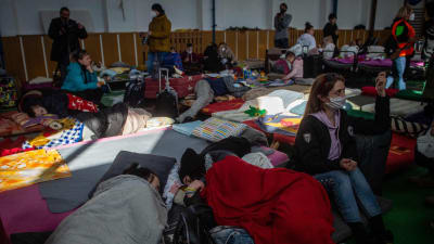 Två personer sover i sovsäckar i en flyktinganläggning. En kvinna sitter bredvid dem.