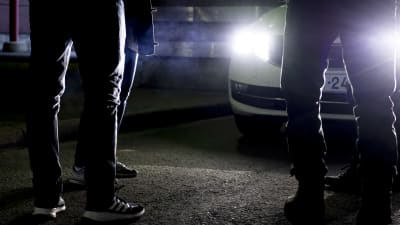 Människors fötter framför en bil i mörkret. Bilen har ljusen tända.
