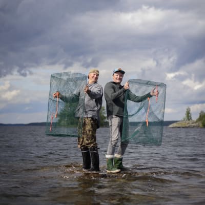 Juha Taskinen ja Kari Hietalahti seisovat kivellä järvessä ja pitävät katiskoja käsissään. 