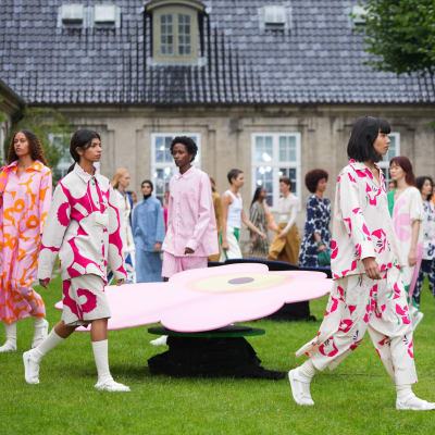 Modeller går på en gräsmatta i Marimekkokläder under en modevisning. I bakgrunden syns ett hus.