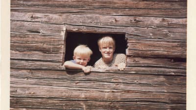 Två leende pojkar tittar ut genom en ojämn fönsteröppning i en stockvägg. 