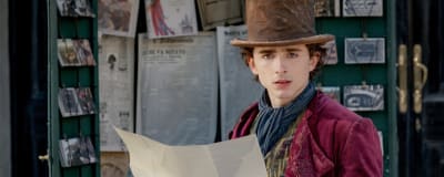Närbild på Wonka (Timothée Chalamet) som har en vinröd jacka, cylinderhatt och står vid ett skyltfönster och håller i ett stort papper.