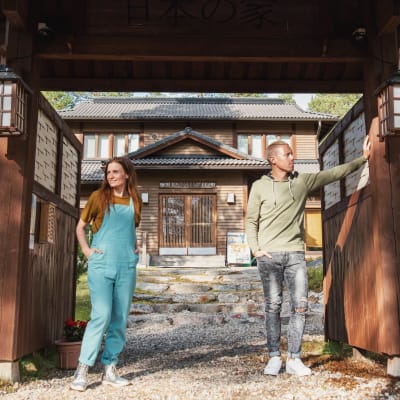 Nainen ja mies seisovat japanilaistyylisellä portilla, taustalla näkyy piha ja talo.