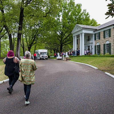 Turister köar mellan de vita pelarna vid ingången till Graceland. Stora träd växer vid uppfarten till byggnaden och i förgrunden går ytterligare ett par turister mot huset.