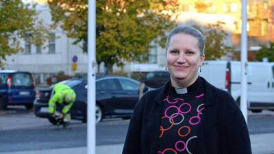 Kira Ertman poserar framför flaggstänger och en man i gul skyddsoverall.