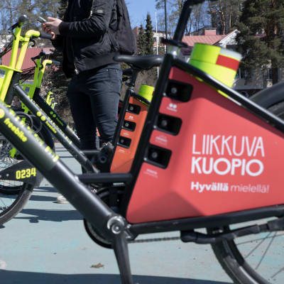 Henkilö parkeeraamassa Vilkku-kaupunkipyörää Kuopion linja-autoasemalla.