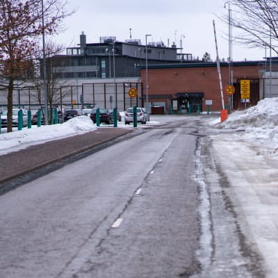Porten till Vanda fängelse, en isig bilväg med en vägbom, bakom den en tegelbyggnad bakom ett högt stängsel.