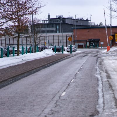 Porten till Vanda fängelse, en isig bilväg med en vägbom, bakom den en tegelbyggnad bakom ett högt stängsel.