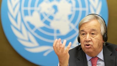 Antonio Guterres med handen i vädret, i bakgrunden FN-logon.