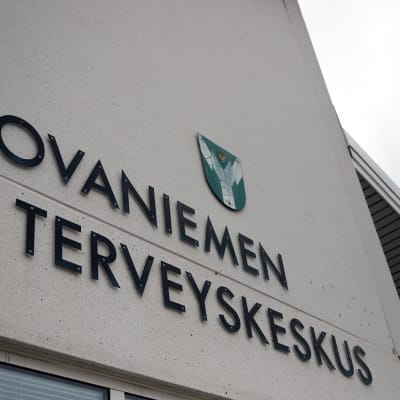Valkoisella ulkoseinällä mustilla kirjaimilla oleva teksti Rovaniemen terveyskeskus ja kaupungin vaakuna.