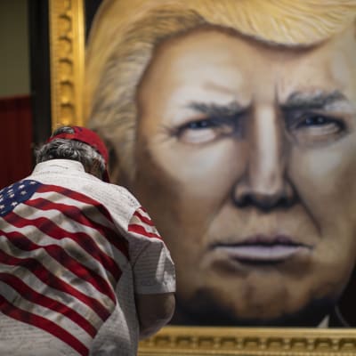 Man i fägrgglad skjorta med USA-flagga beundrar tavla av Donald Trump.