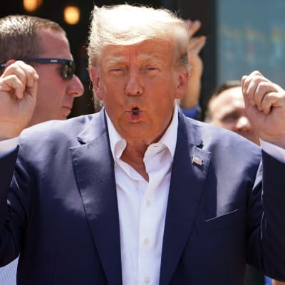 Donald Trump putar med läpparna och höjer båda händerna i en entusiastisk gest. Han är klädd i blå kavaj, vit skjorta, utan slips och med en amerikansk flagga på kavajuppslaget.