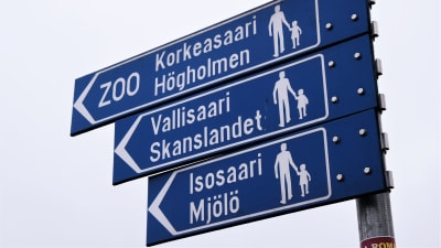 En skylt på vilken det står att man ska gå till vänster för att komma till Mjölö. Skylten är blå, formad som en pil, och texten är vit. Bakgrunden är molnig.